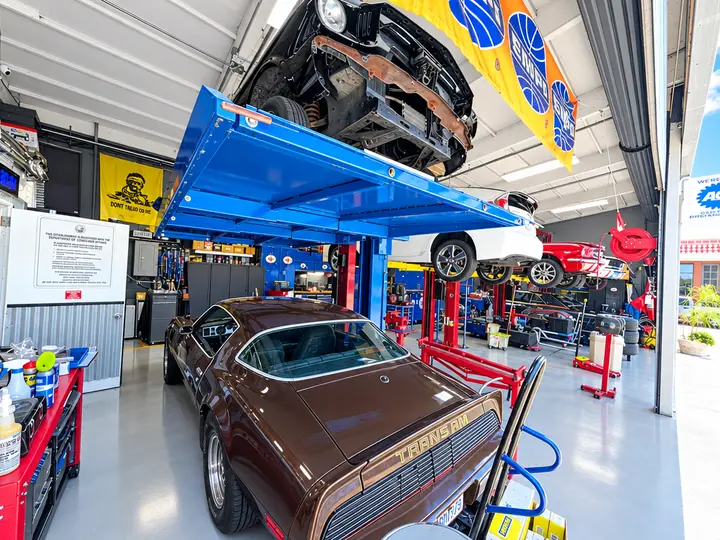 Brown Pontiac Trans Am under a project car on a four-post lift at Cedillo's classic auto repair in Fullerton
